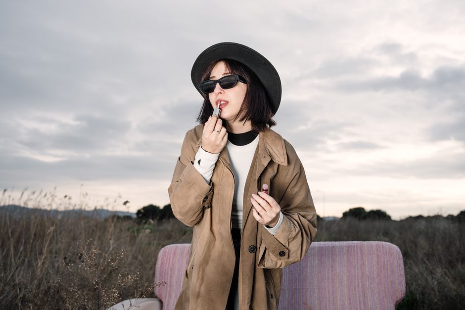 A fashionable woman in a trench coat and hat applies lipstick in a natural setting.