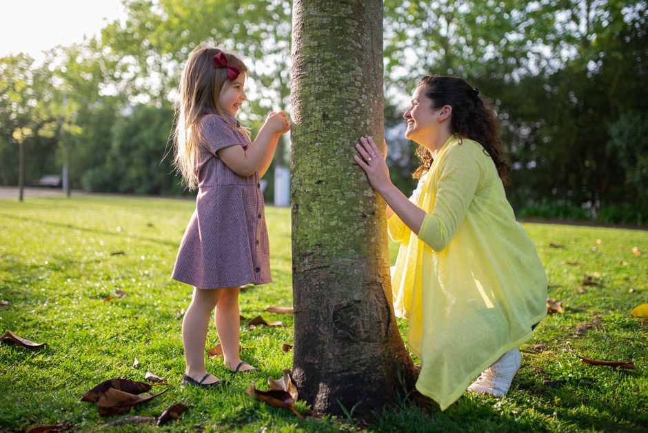 A joyful mother and daughter playing hide and seek around a tree in a sunny park setting.