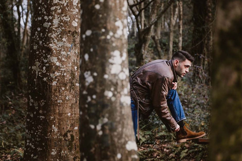 A man in leather jacket tying brown boots in a wooded area. Captured outdoors.