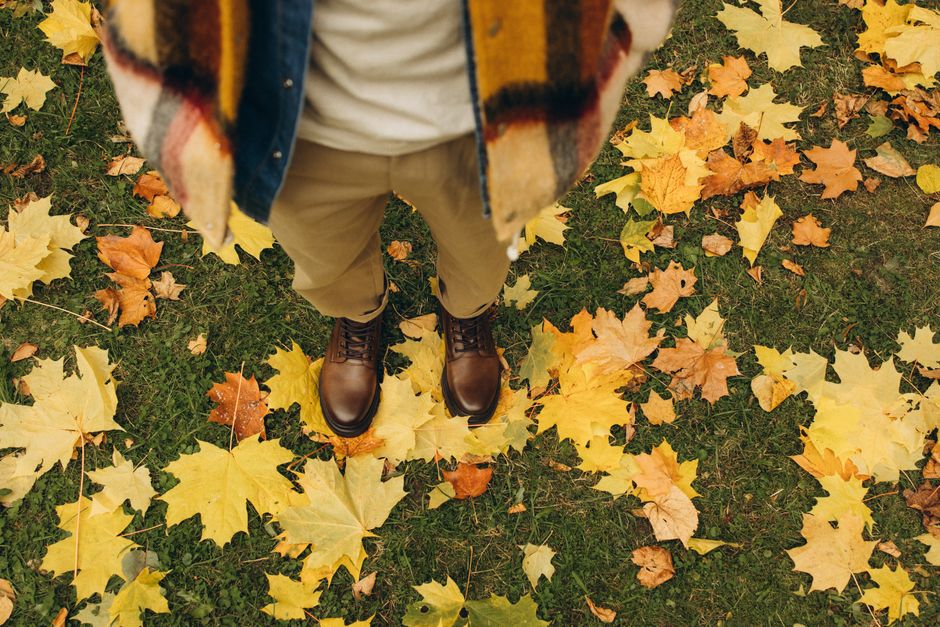 A person stands on autumn leaves wearing brown boots and a cozy scarf.