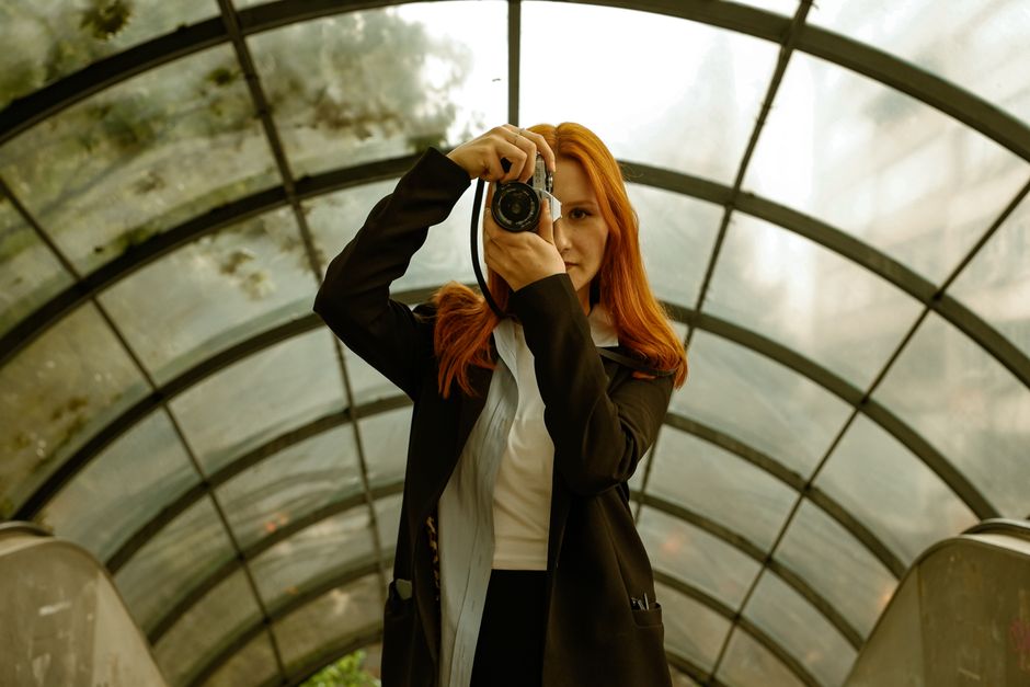 A redhead woman holding a camera inside a curved glass tunnel, creating a unique perspecti
