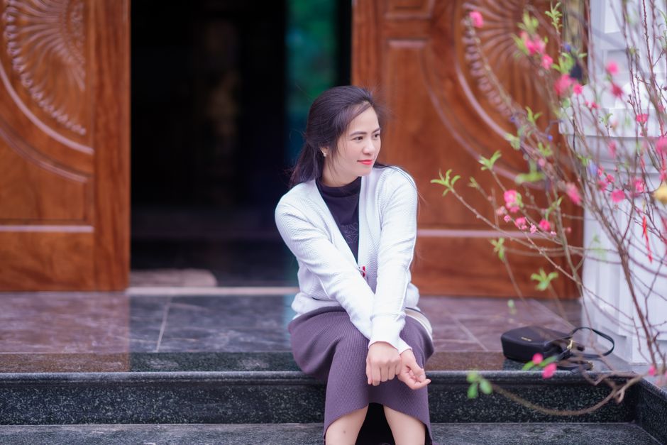Asian woman sits at a traditional temple entrance, surrounded by blooming pink flowers.