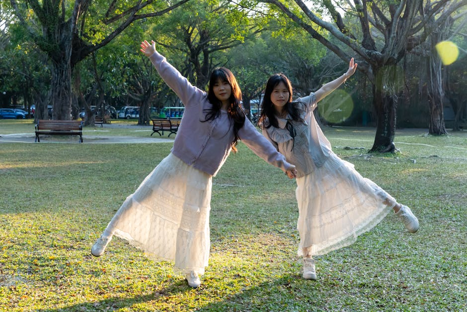 Two people posing playfully in a sunlit park setting surrounded by lush trees.