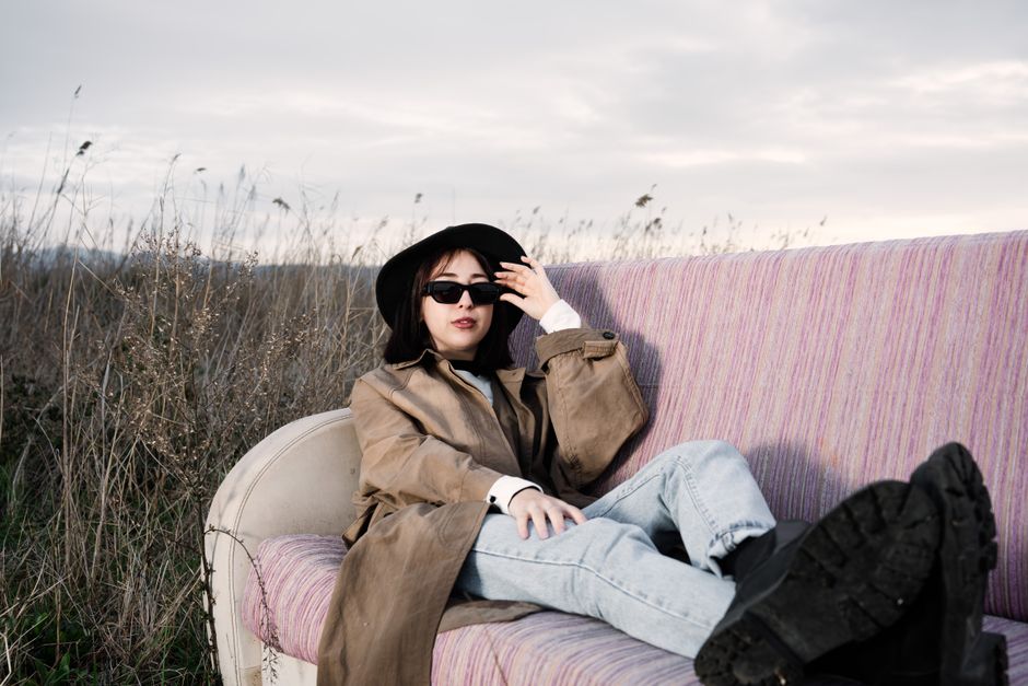 Woman in a trench coat and sunglasses lounging on a sofa outdoors.