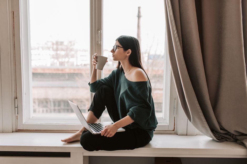 Side view full length of focused young lady in eyeglasses and casual outfit sitting on windowsill with legs crossed and browsing laptop while drinking hot beverage and looking away pensively