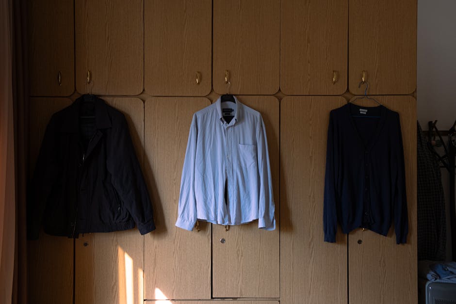 Wardrobe with clothes hanging, featuring a black jacket, white shirt, and dark sweater. Indoor setting.