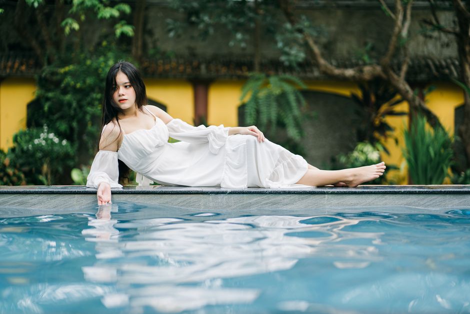 Full body of serious Asian female in long dress looking away while lying on edge of swimming pool near building