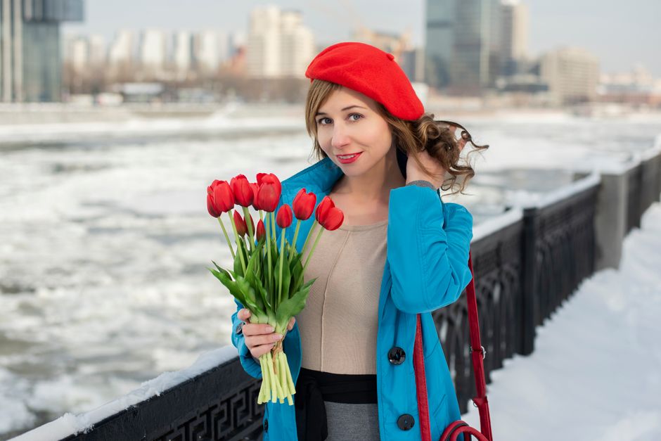Woman in urban winter scene holding red tulips, dressed in blue coat and red beret.