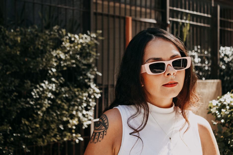 Confident woman wearing sunglasses and displaying tattoos in a sunny outdoor setting.