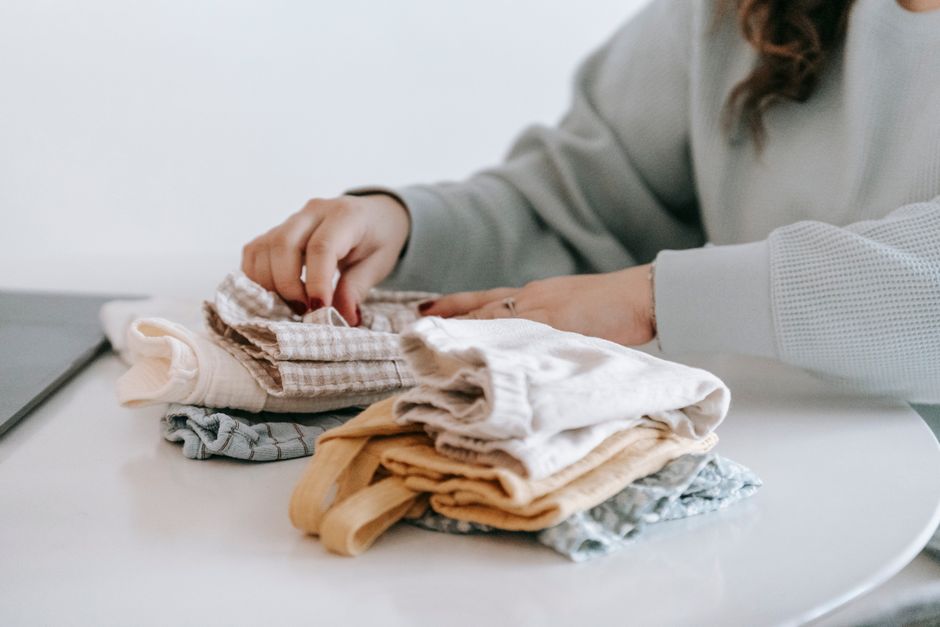 Crop faceless female in casual clothes folded colorful baby garments while sitting at whit