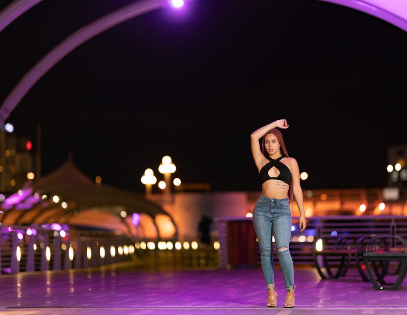 Stylish woman in black top and denim posing at night on a brightly lit urban bridge.