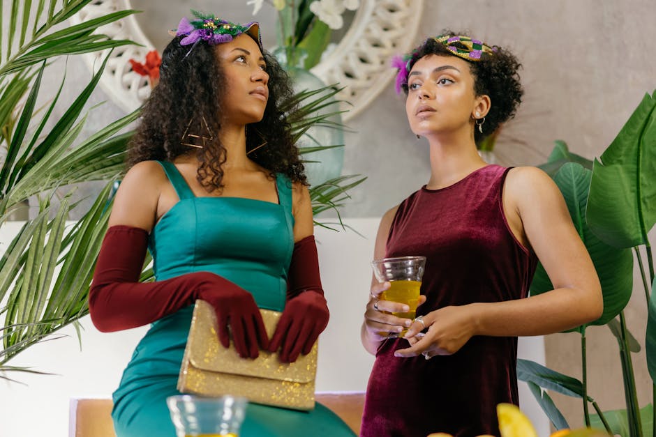 Two women in stylish dresses and masks enjoying a glamorous masquerade party with drinks.