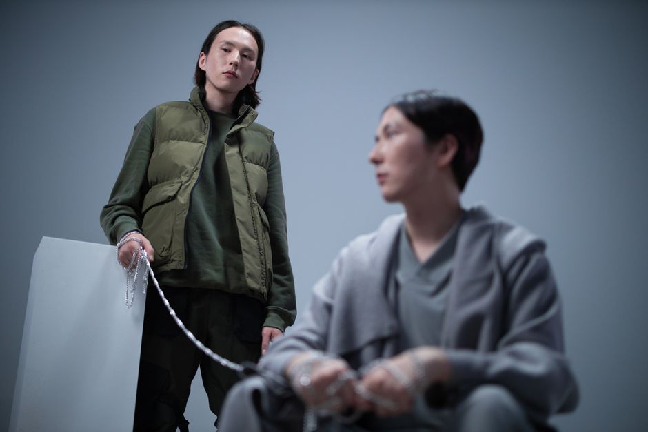 Two men in modern outerwear posing in a studio with chains on a grey background.