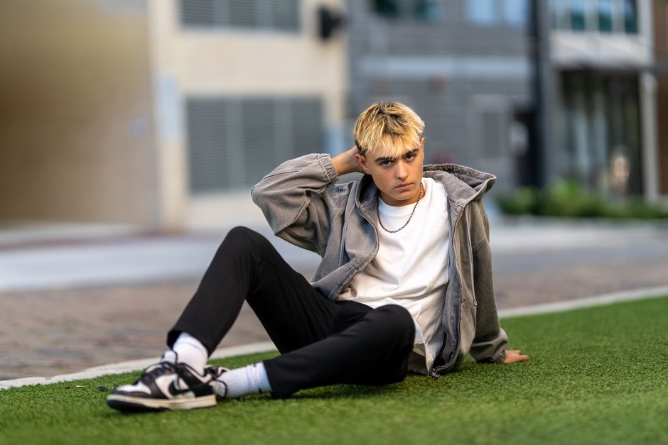 Casually dressed young man sits on grass in an urban environment.