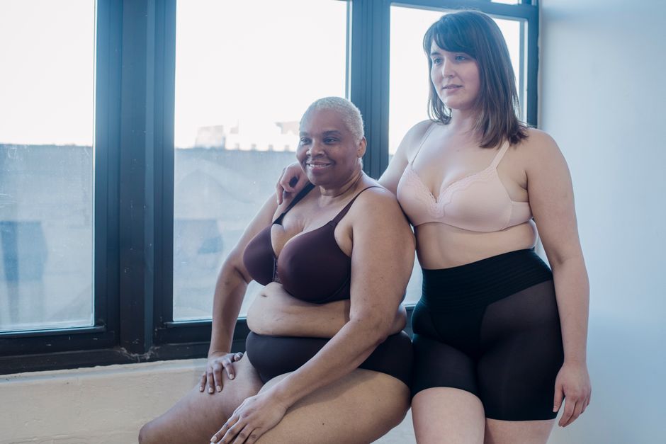 Overweight woman in lingerie embracing cheerful African American model while looking forward in daytime indoors