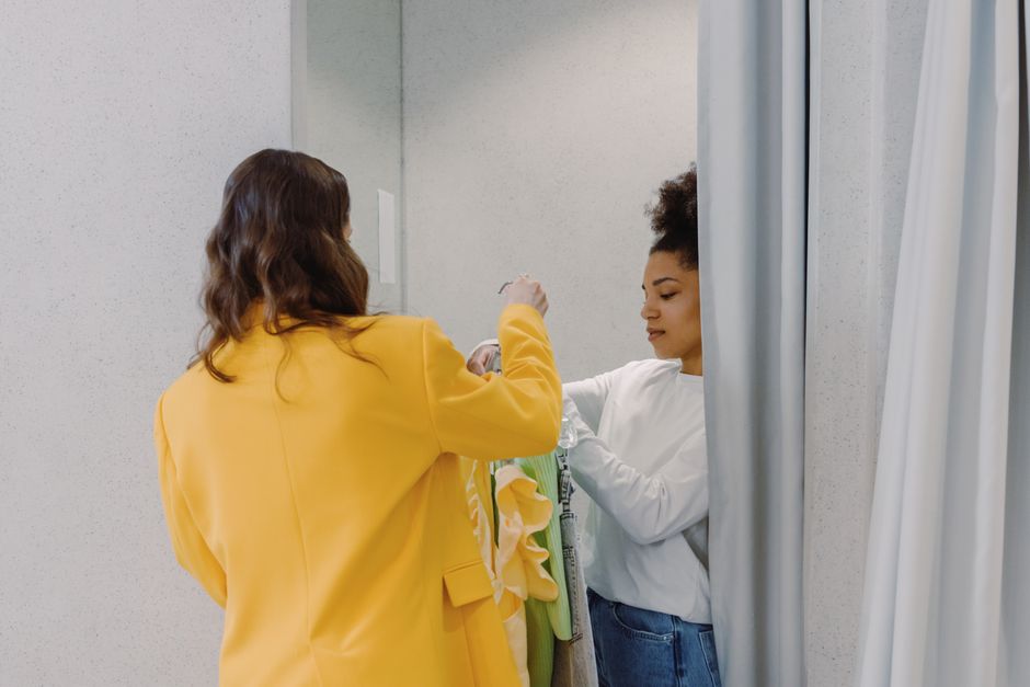 A personal stylist helps a client with clothing choices in a fitting room, focusing on fashion styling.