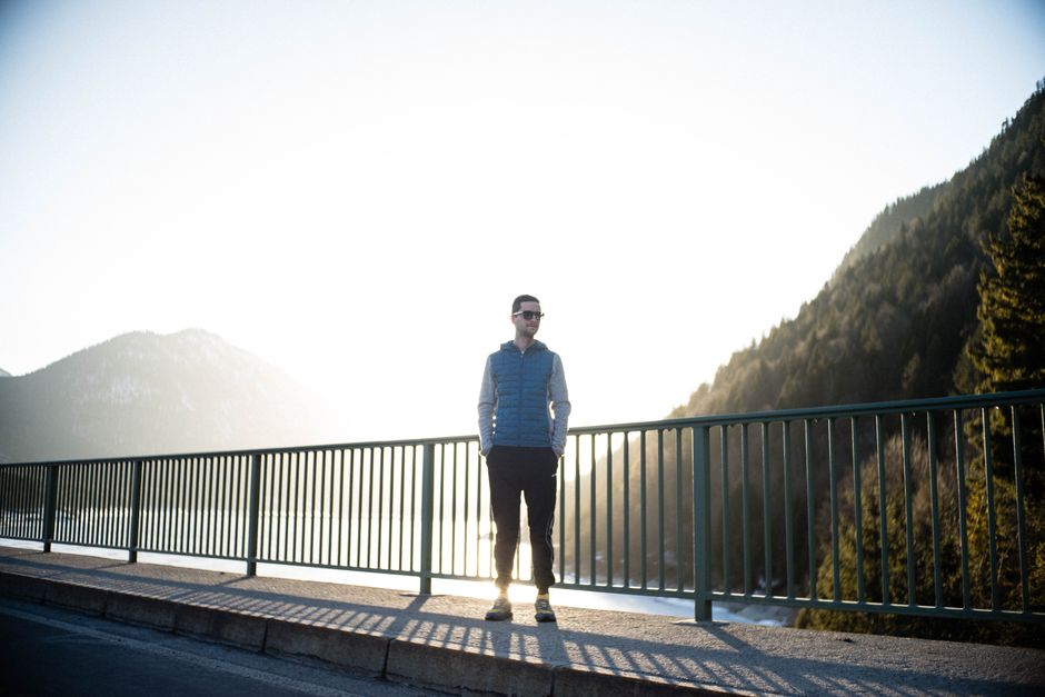 A man stands on a bridge in Lenggries, Germany during sunset, surrounded by mountains.