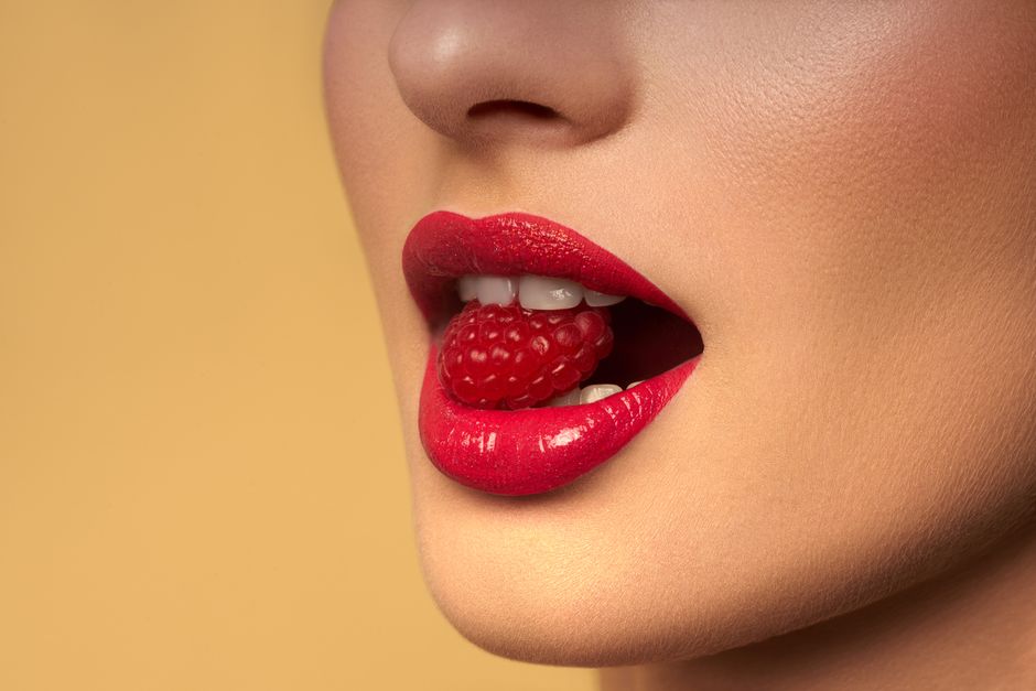 Vibrant red lips holding a fresh raspberry in sensual close-up shot.