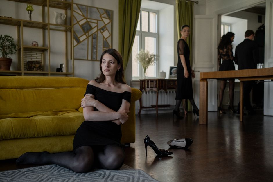 A woman in a black dress sits thoughtfully indoors during a stylish social gathering.