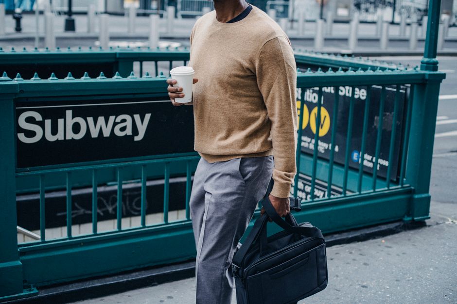 Side view of African American male strolling near green barrier of subway entrance with cup of coffee and black briefcase