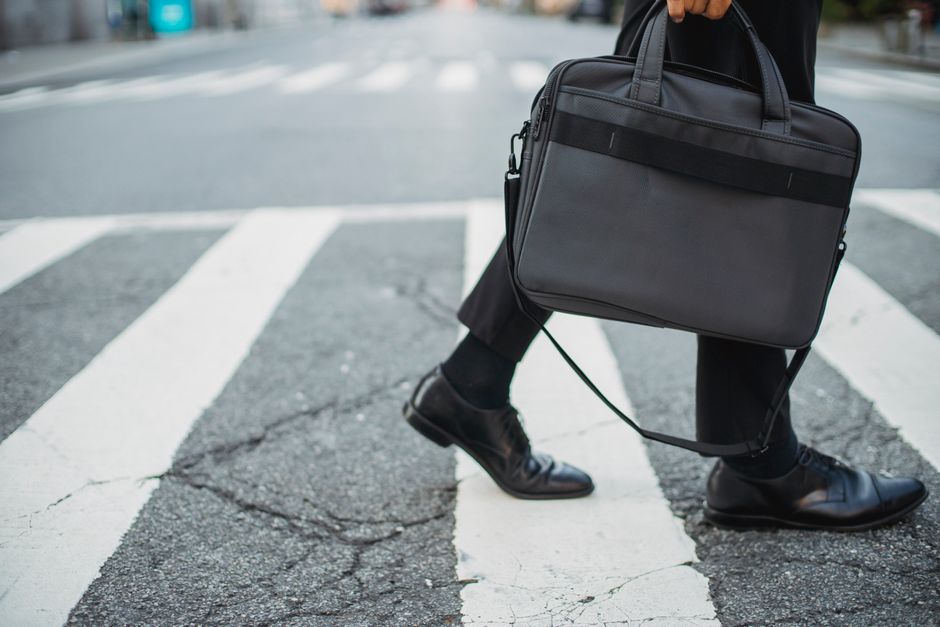 A business professional in leather shoes crosses a city pedestrian lane with a black bag.
