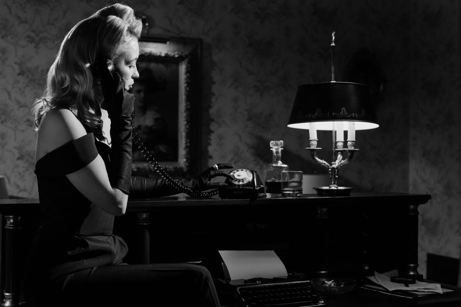 Black and white portrait of an elegant woman in vintage attire, using a retro telephone in a dimly-lit room.