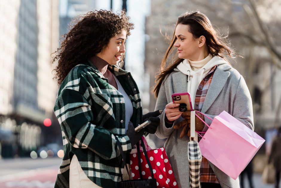 Young women in warm clothes with paper bags standing on city street using cellphones and having conversation while looking at each other in daylight