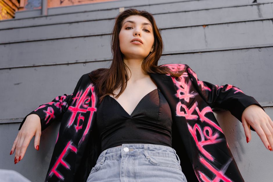 Confident woman in a black blazer with pink graffiti design, leaning back on steps in an urban setting.