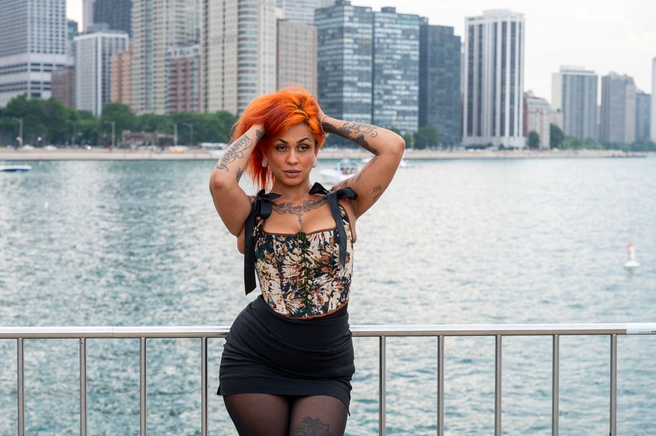 Stylish woman with orange hair poses by a city waterfront with urban buildings in the background.
