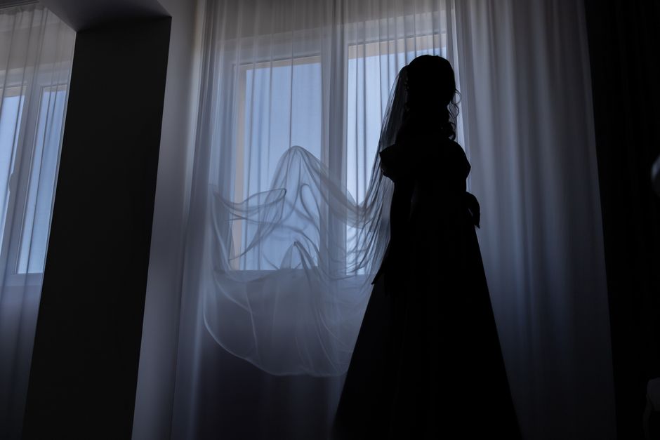 A mysterious silhouette of a bride stands by a window with her veil flowing gently.