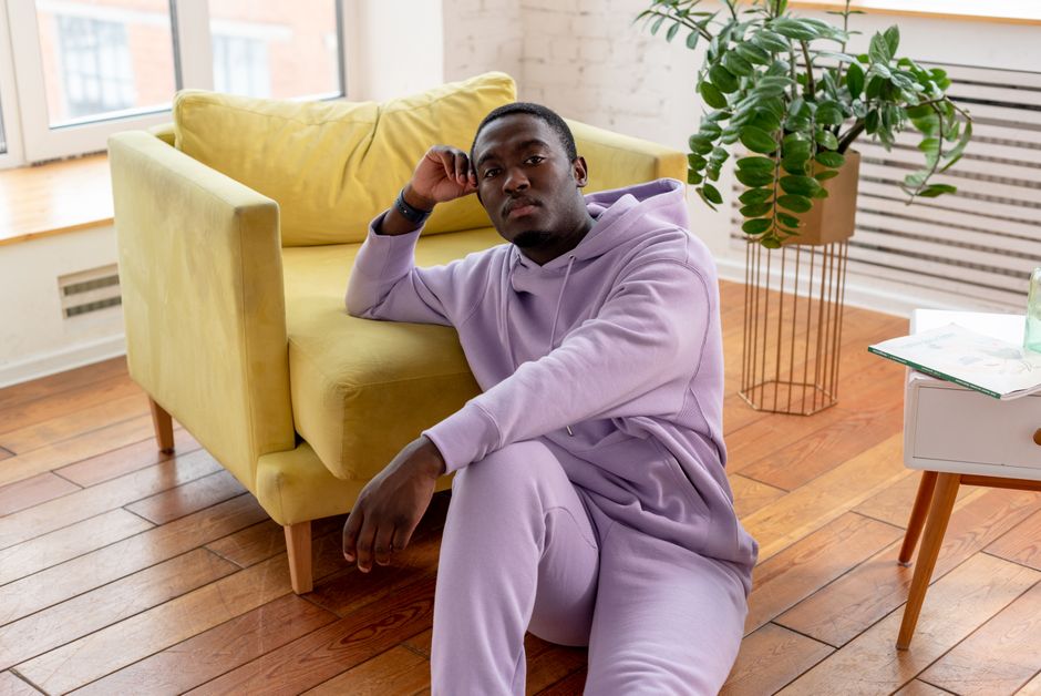 Pensive African American with short dark hair in trendy clothes sitting on floor and leaning on yellow armchair in light living room in daytime