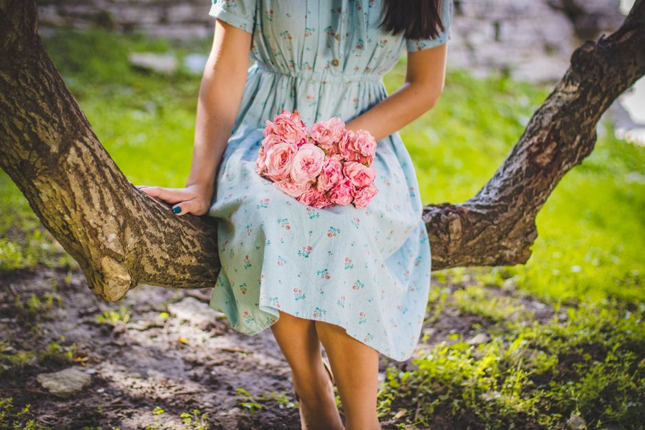 Young woman holding pink flower bouquet while sitting on a tree branch outdoors.