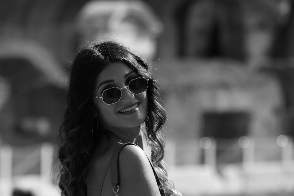 Black and white image of a woman smiling while wearing sunglasses, captured outdoors.