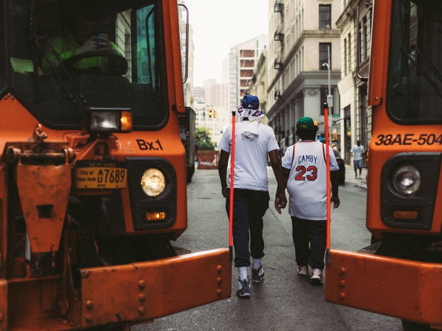 Two people walk between orange New York City street trucks in an urban setting.