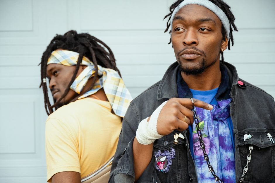Cool African American men with dreadlocks in casual wear and fabric headbands standing back to back next to grey wall
