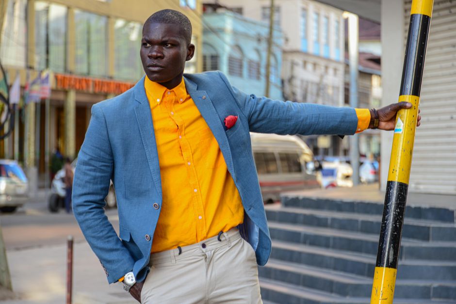 Stylish man in a blue coat and yellow shirt posing with confidence in an urban setting.