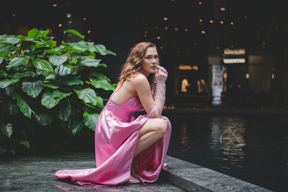 Stylish woman in a pink dress poses elegantly by a reflective water pool, surrounded by lush greenery.