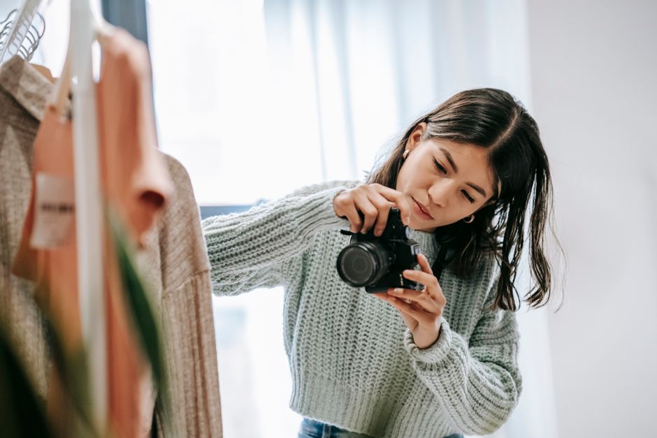 Young concentrated female photographer taking picture of clothes rack with hanging garment while looking at camera display in light room